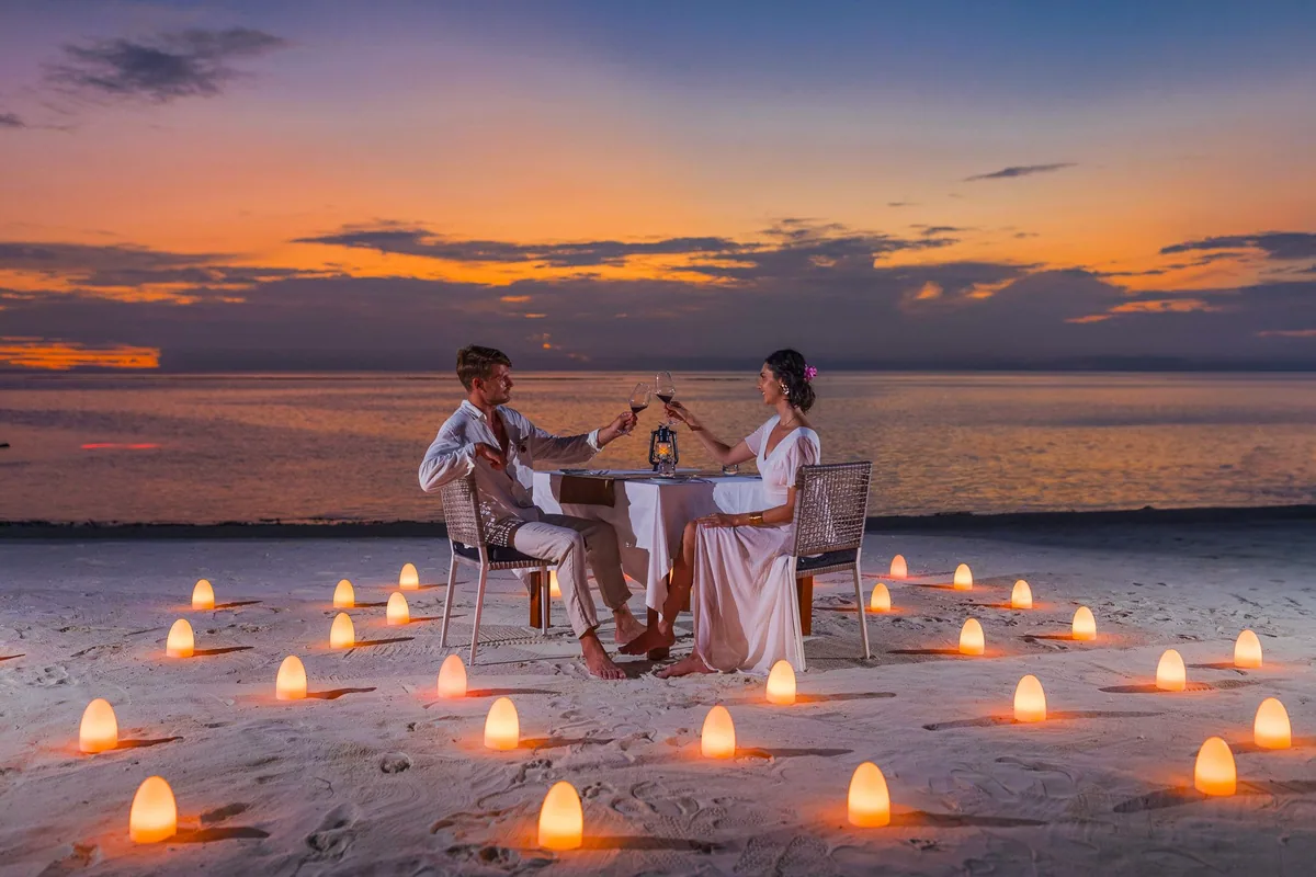 VARU by Atmosphere, lifestyle, North Malé Atoll, Maldives, Sunset destination-dining setup on the beach, a couple toasting wine at a small white-linen table at sunset, ringed by a perfect circle of glowing wax candles set into the sand, with the silhouetted ocean horizon and pink-orange dusk sky behind.