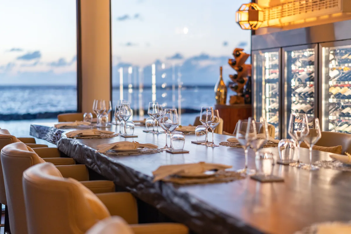 VARU by Atmosphere, dining, North Malé Atoll, Maldives, NU Cellar over-water wine room interior at sunset, the long live-edge wooden table with sand-coloured leather club chairs, twin wall-mounted glass-fronted wine fridges with vintage display, the floor-to-ceiling window opening onto the open ocean and pale sunset sky.