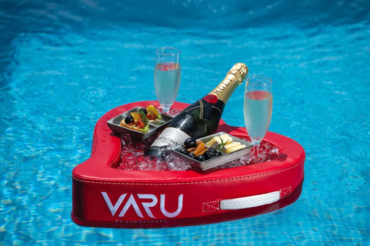 VARU by Atmosphere, lifestyle, North Malé Atoll, Maldives, In-pool floating breakfast service, a red heart-shaped float with the embossed VARU BY ATMOSPHERE wordmark, a bottle of Moët champagne in the ice well, two tasting plates of canapés and cheese, and two flute glasses, on the rippled blue pool surface.