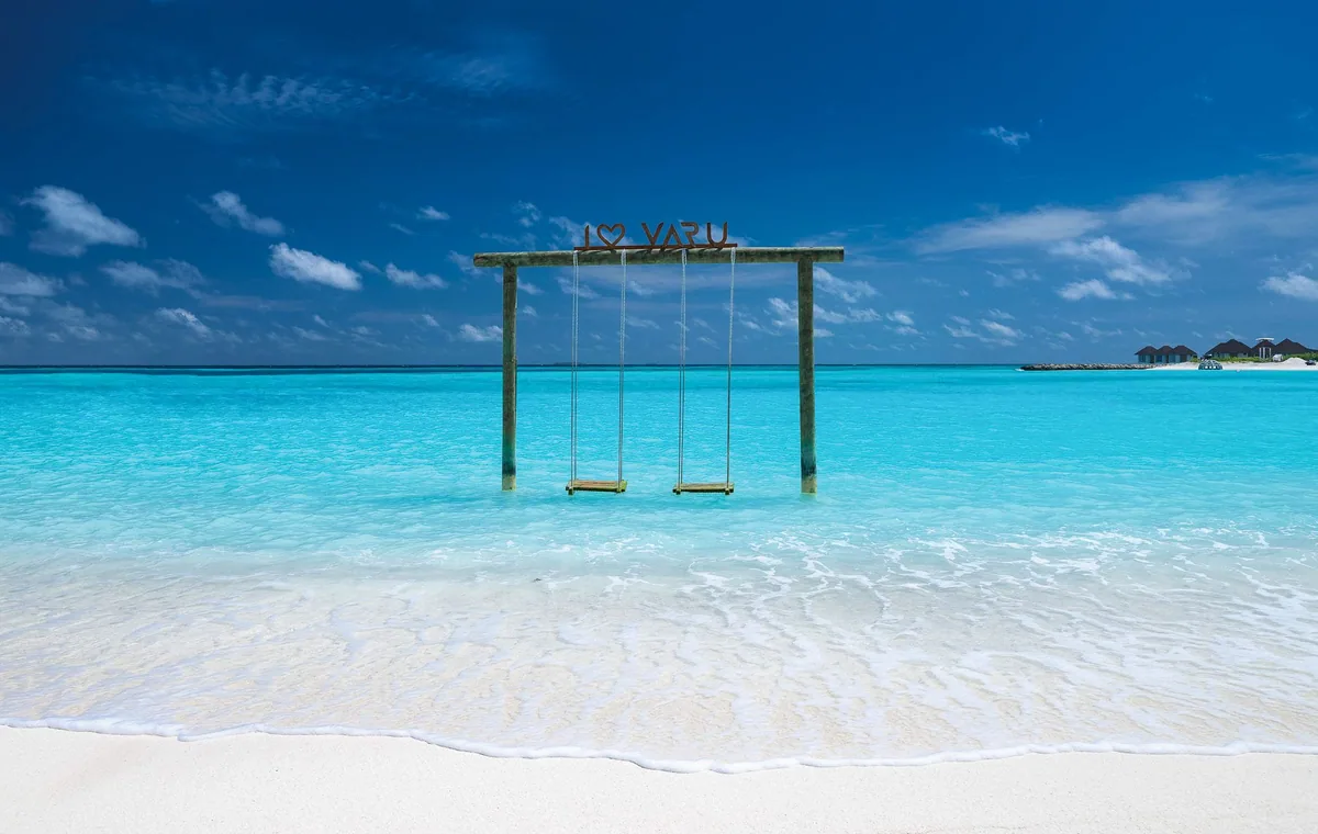 VARU by Atmosphere, lifestyle, North Malé Atoll, Maldives, The signature I LOVE VARU beach swing, the standalone wooden swing-frame set in the calm shallow lagoon with the wordmark mounted at the crossbeam, two timber plank swings dipping just above the water, and the perimeter overwater villa cluster visible at the distant right edge of the property.