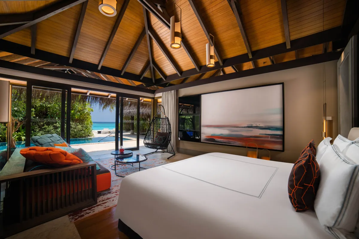 Velaa Private Island, villa beach, Noonu Atoll, Maldives, Beach Pool Villa bedroom (alternate angle), daybed with orange and teal cushions, hanging black-rattan chair, abstract orange-blue painting above the headboard, glass doors open to the private pool, beach, and lagoon beyond.