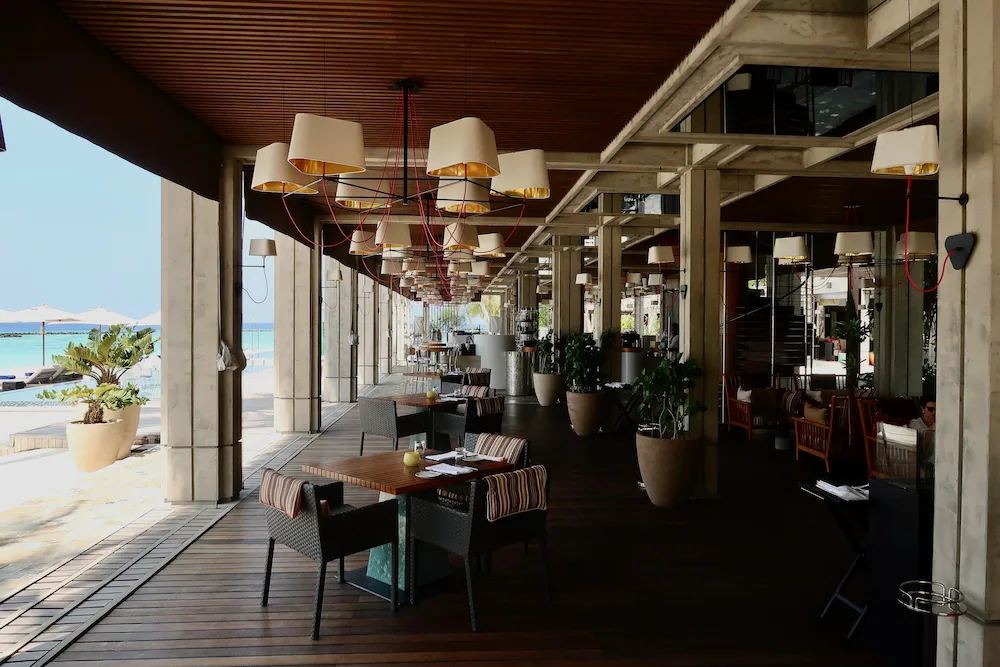 Velaa Private Island, dining, Noonu Atoll, Maldives, Athiri's all-day open-air pavilion: exposed concrete columns supporting a low timber ceiling, drum-shade pendants on red wiring, beachfront banquette seating with striped fabric, banana plants in stone planters.