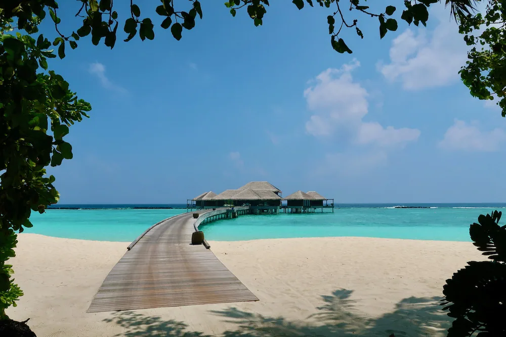 Velaa Private Island, spa, Noonu Atoll, Maldives, The Velaa spa complex at the end of a curved timber boardwalk crossing the lagoon from a white-sand beach, thatched-roof treatment pavilions on stilts over turquoise water, framed by tropical foliage.