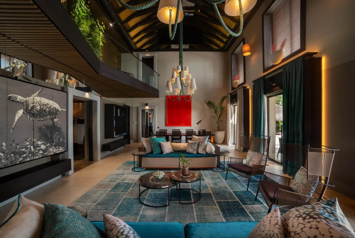 Velaa Private Island, villa residence, Noonu Atoll, Maldives, Romantic Pool Residence living room: double-height vaulted ceiling with a green-leaf installation suspended from the rafters, custom multi-shade chandelier, a large turtle photo work and a red Rothko-style canvas on the back wall, teal patchwork rug, sculptural seating around marble coffee tables.