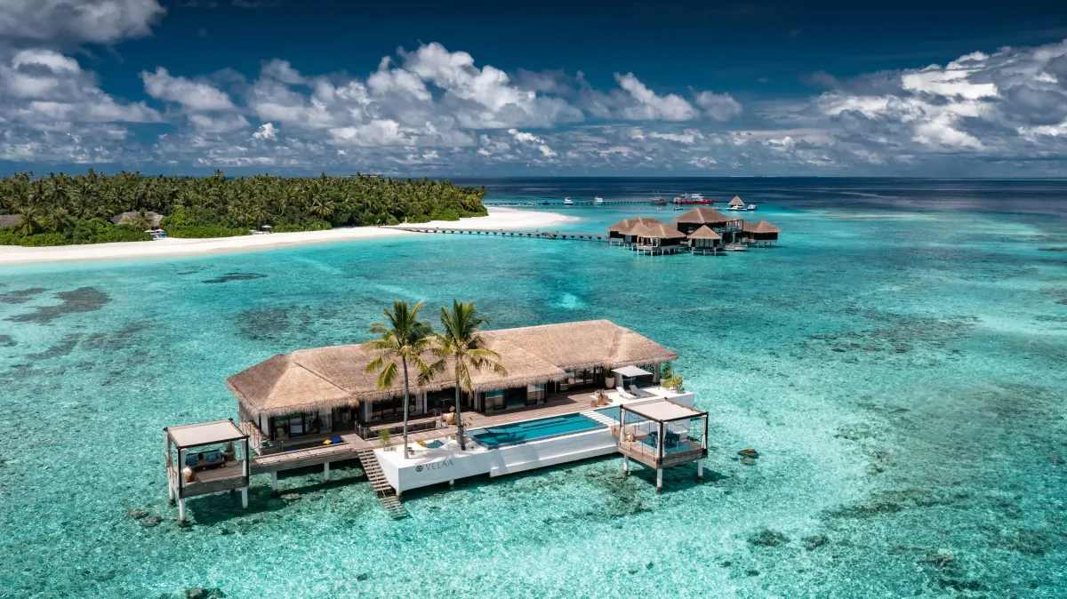 Velaa Private Island, villa overwater, Noonu Atoll, Maldives, Aerial of the Velaa overwater villa cluster, thatched-roof bungalow with cantilevered private-pool deck over turquoise lagoon, second overwater pavilion to the right, beach point and palm-covered island edge in the background.