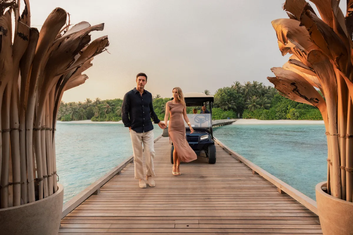 Velaa Private Island, lifestyle, Noonu Atoll, Maldives, Arrival on the timber boardwalk, the Velaa-signature dried palm-trunk sculptural planters flanking the path, electric golf cart standing by, the lagoon and main island foliage in the background.