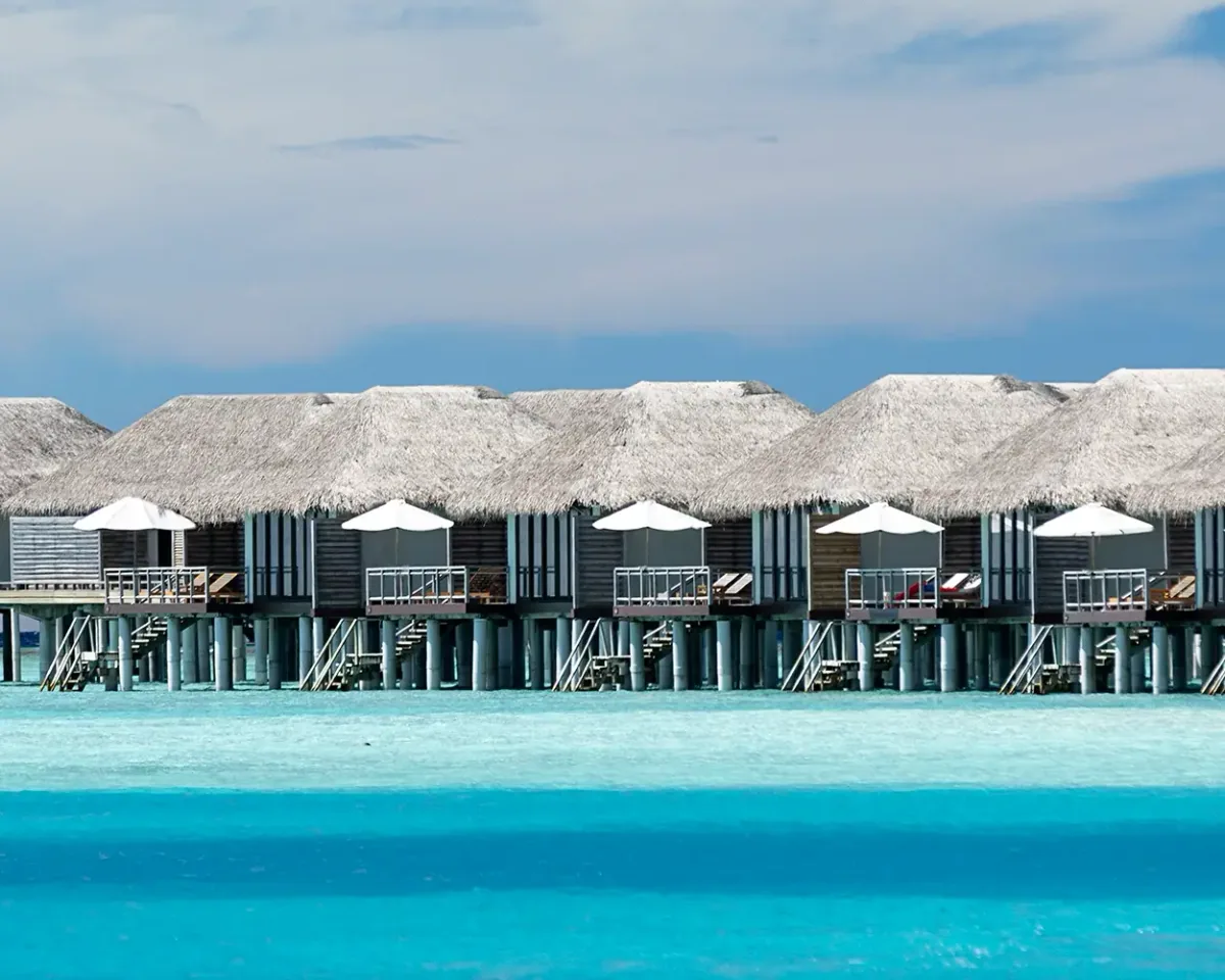 Velassaru Maldives, villa overwater, South Malé Atoll, Maldives, Velassaru water villas row at the over-water configuration: thatch-roof villas on stilts with white parasols on each deck, sun loungers, and vivid turquoise lagoon edge running the length of the South Malé Atoll cluster.