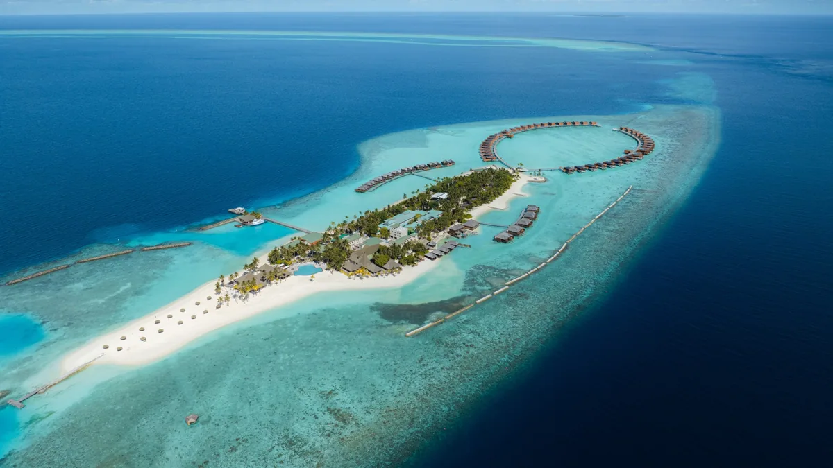 Veligandu Island Resort & Spa, aerial, Alif Alif Atoll (North Ari, Rasdhoo cluster), Maldives, Veligandu sandbank-extension aerial, the elongated natural island with the country-distinctive sandbank trailing into the lagoon at the northern tip, overwater villa ring wrapping the southern lagoon edge, surrounding Rasdhoo cluster reefs visible. The property-name signature shot (Veli + gandu = sand-tongue).