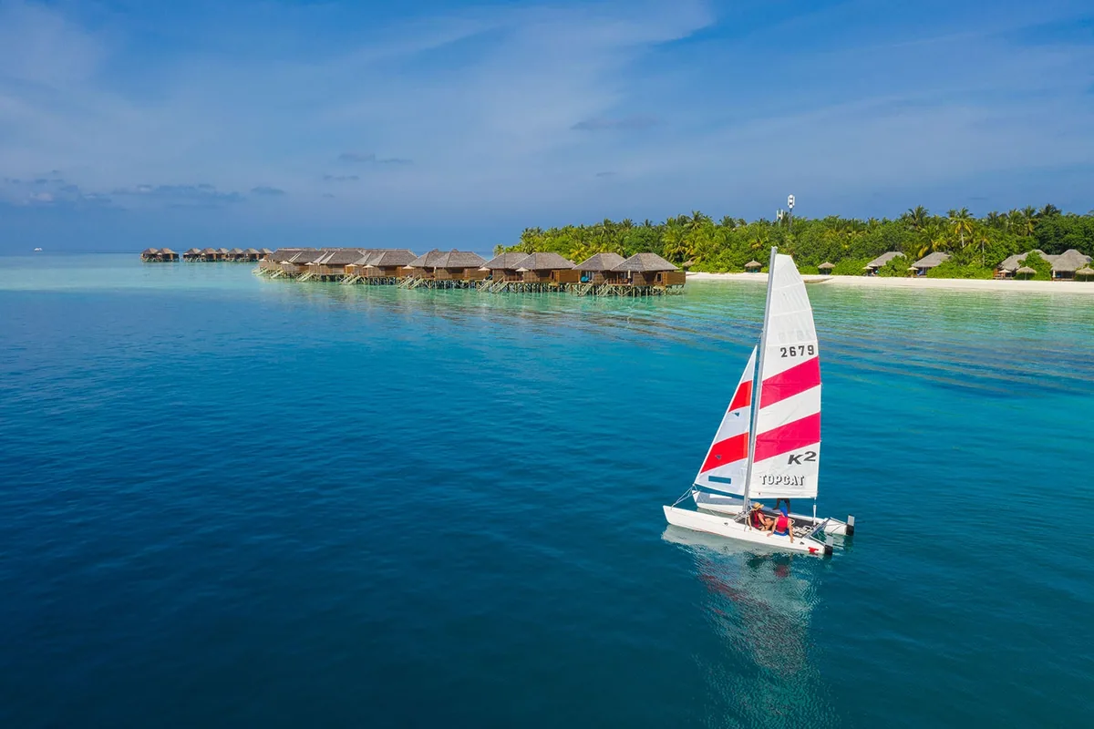 Veligandu Island Resort & Spa, activities, Alif Alif Atoll (North Ari, Rasdhoo cluster), Maldives, Topcat catamaran (number 2679, red-and-white striped sail) sailing across the deep-blue Veligandu lagoon, the overwater villa row and main island reading along the coast in the distance. The property's sailing programme as part of the watersports cluster.