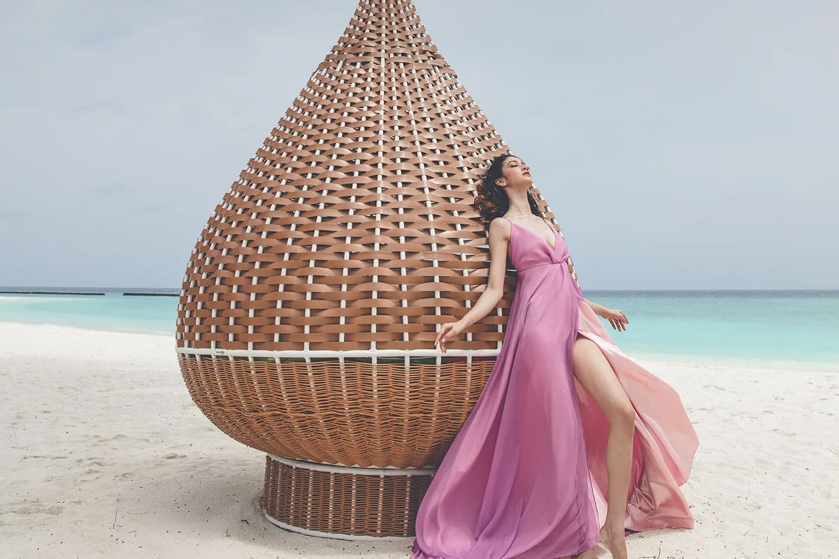 Veligandu Island Resort & Spa, setting, Alif Alif Atoll (North Ari, Rasdhoo cluster), Maldives, Teardrop-shaped woven-rattan beach pavilion sculpture on the white sand, a model in a flowing pink dress posed against its lattice-weave structure, the turquoise lagoon and reef-line in the background. Veligandu's signature beach-art installation.