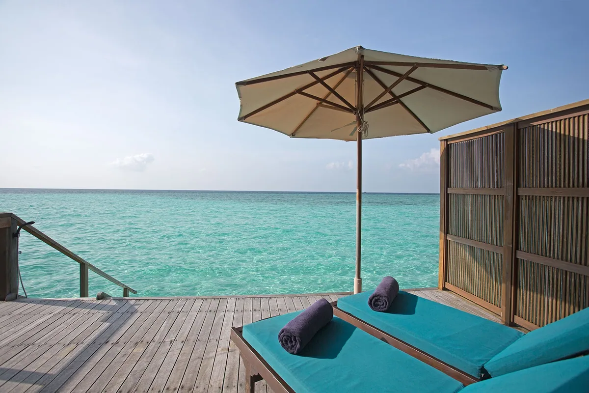 Veligandu Island Resort & Spa, villa overwater, Alif Alif Atoll (North Ari, Rasdhoo cluster), Maldives, Sunset Jacuzzi Water Villa deck, twin turquoise-cushioned sun-loungers under a wood-frame canvas umbrella on the timber deck, woven privacy-screen panel at the side, deck steps descending into the deep-blue lagoon facing the sunset axis.
