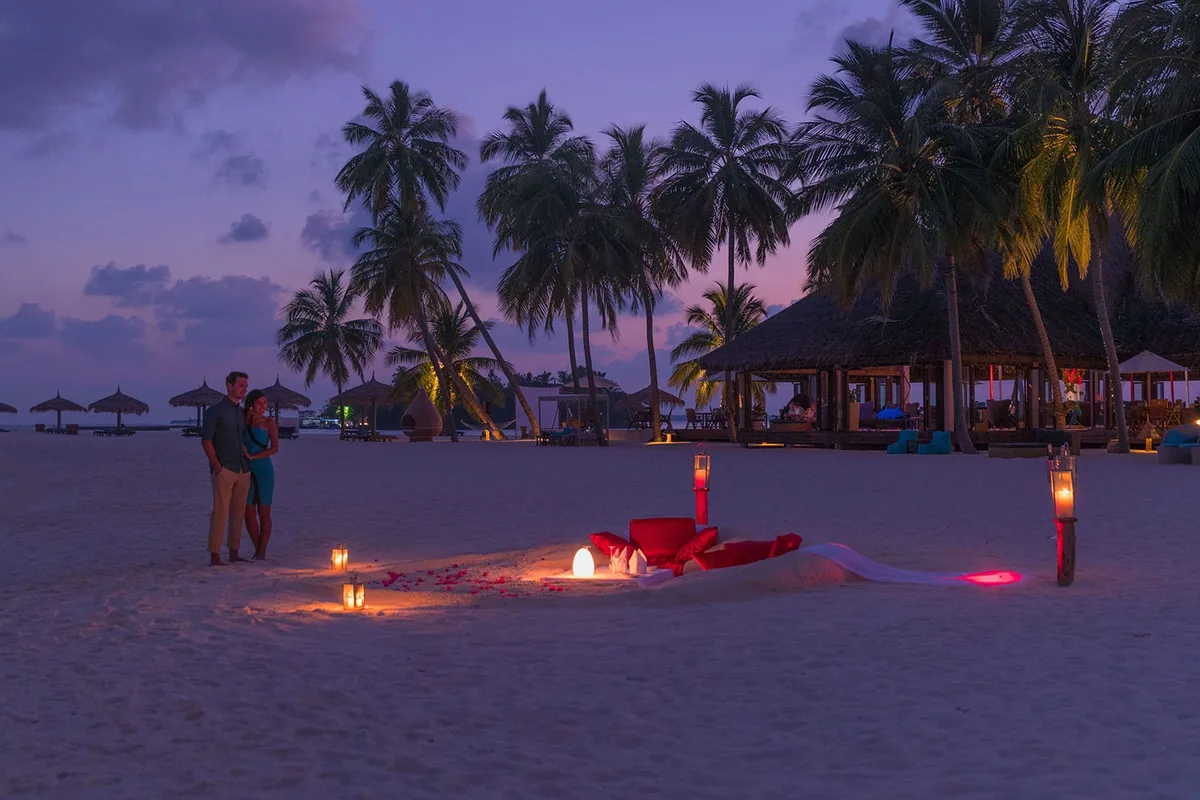 Veligandu Island Resort & Spa, dining, Alif Alif Atoll (North Ari, Rasdhoo cluster), Maldives, Romantic beach dining at twilight, a couple standing beside the candle-lit table setup on the white sand with red rose-petal letters laid out at their feet, hurricane-glass candles and palm canopy framing, the principal dining pavilion lit warmly in the background.