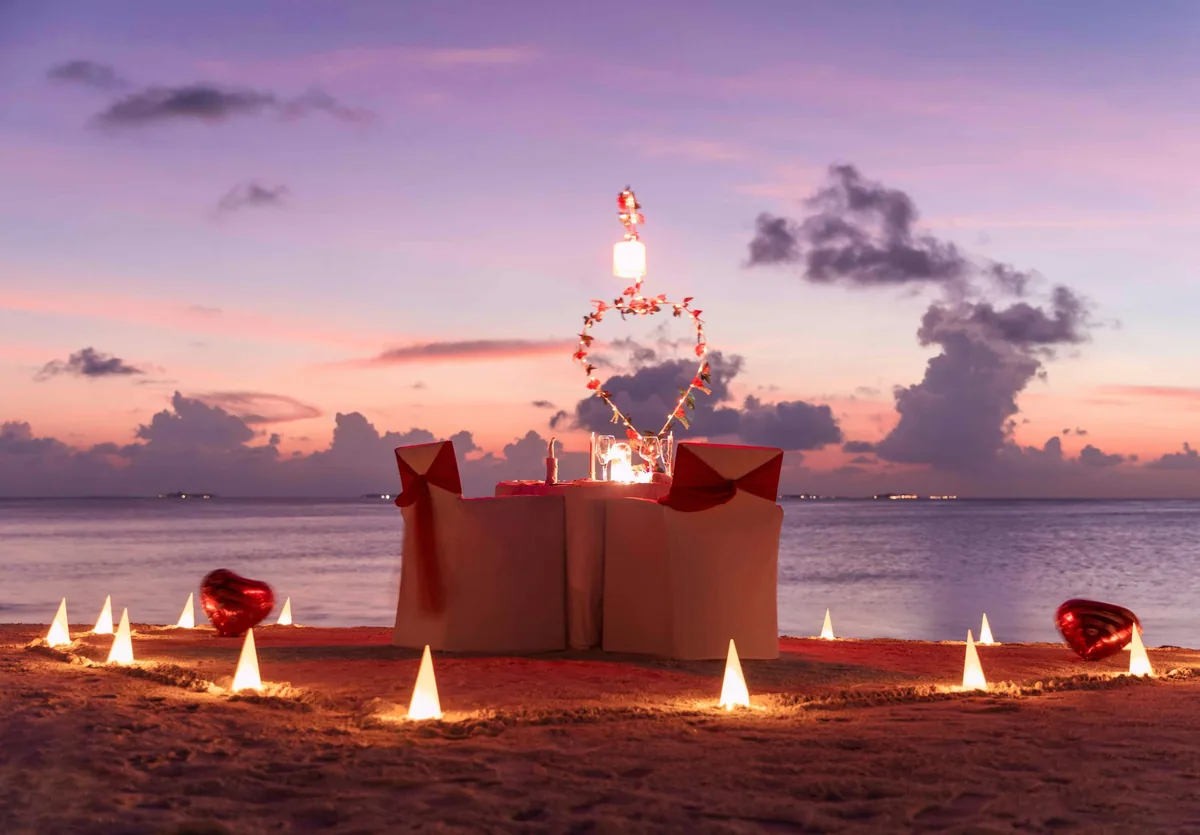 Vilamendhoo Island Resort & Spa, dining, Alif Dhaal (South Ari) Atoll, Maldives, Private Valentine's beach dinner setup with red-bow-tied chair covers, a heart-shaped fairy-light archway carrying a single lantern, conical white sand-candles outlining a heart on the sand, and red heart-balloons under the pastel pink-and-lavender twilight sky.