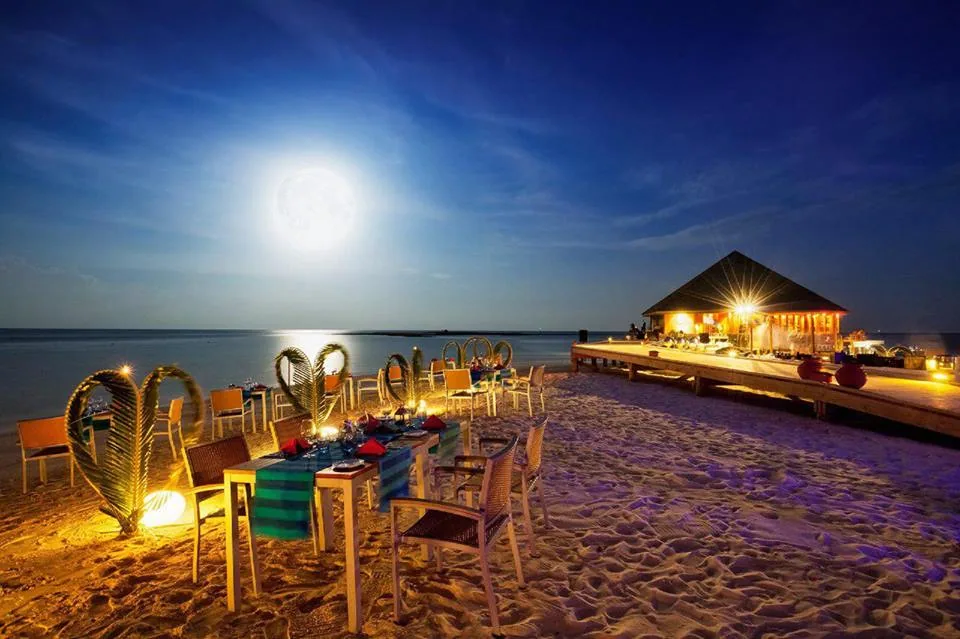 Vilamendhoo Island Resort & Spa, dining, Alif Dhaal (South Ari) Atoll, Maldives, Starlit beach dinner under the full moon over the lagoon, with green-striped table runners on the sand, palm-frond heart silhouettes at the chair backs, candle-lit dining area, and the lit overwater dining pavilion at the right edge.