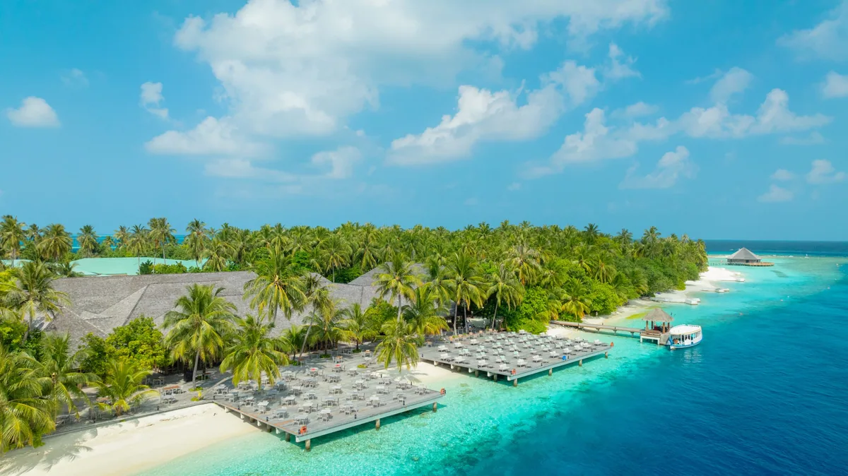 Vilamendhoo Island Resort & Spa, setting, Alif Dhaal (South Ari) Atoll, Maldives, Vilamendhoo jetty aerial with the long timber walkway parallel to the western beach lined with sunbed-and-umbrella pairs, the overwater dhoni-style restaurant pavilion at the boardwalk far end, and the yellow excursion dhoni moored at the jetty mid-frame.