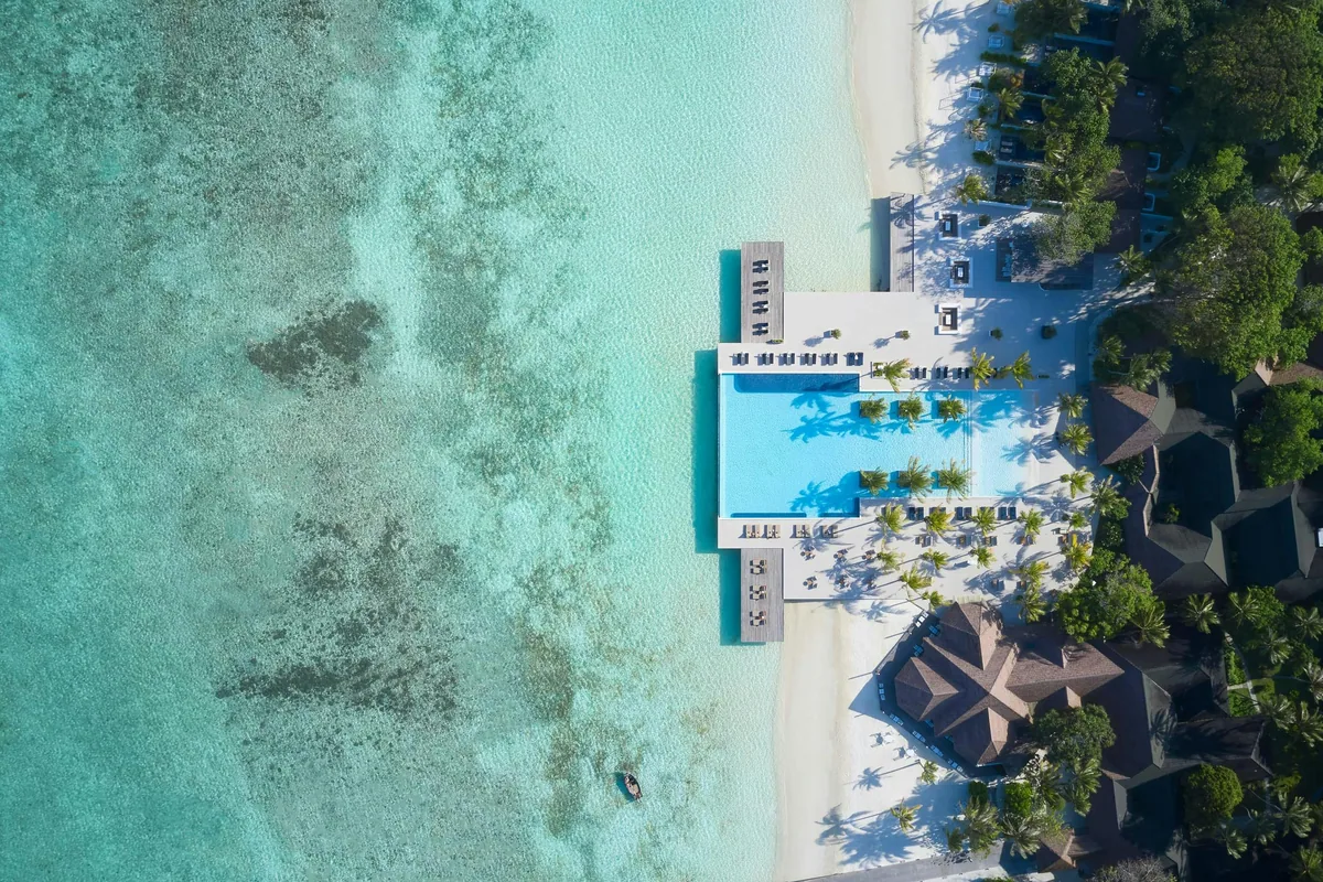 Villa Nautica Paradise Island Resort, aerial, North Malé Atoll, Maldives, Main pool top-down drone aerial: the rectangular pool flush with the edge of the turquoise lagoon, central row of palm-canopied loungers in the water, palm strips flanking the deck on both sides, beach and shallow reef at the lagoon side (Villa Nautica signature pool-deck shot).