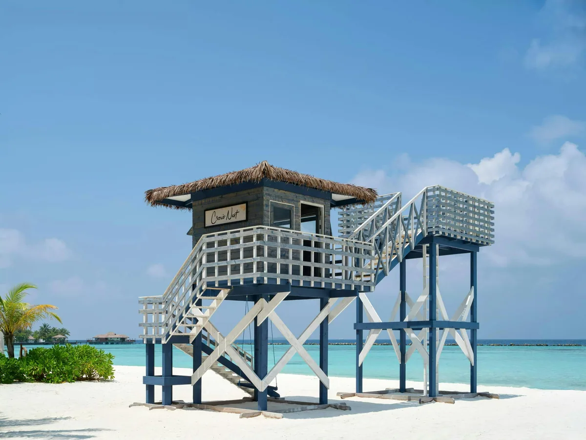 Villa Nautica Paradise Island Resort, setting, North Malé Atoll, Maldives, Crow's Nest beach lookout: elevated lattice-walled tower with thatched roof, navy-and-white painted nautical structure on white sand, deck stairs up to the cabin platform, palm cluster at left, turquoise lagoon and overwater villa silhouettes at horizon.