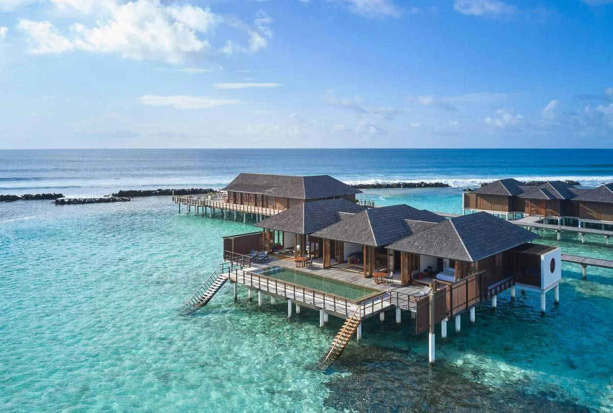 Villa Nautica Paradise Island Resort, villa overwater, North Malé Atoll, Maldives, Two-Bedroom Ocean Suite with Pool aerial: multi-pavilion overwater suite with shingled hip roofs, central deck with infinity plunge pool spilling toward the lagoon, steps descending into clear shallows, breakwater visible in the distance, adjacent water villa cluster on the right.