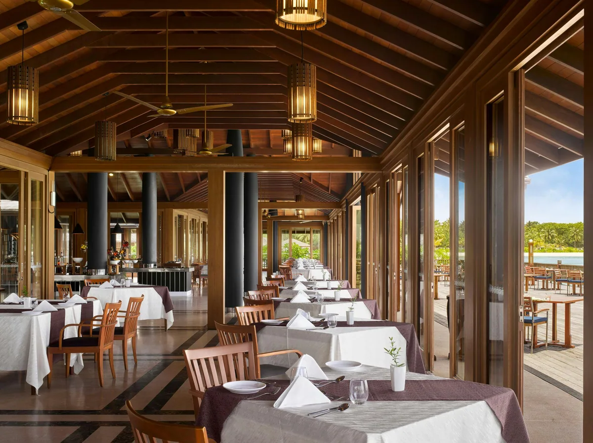 Villa Nautica Paradise Island Resort, dining, North Malé Atoll, Maldives, Lagoon Restaurant interior: dark-wood vaulted pavilion with exposed beam ceiling, brass hanging lanterns, ceiling fan, white-clothed tables down both sides with caned-back chairs, large open windows to the deck and lagoon at right.