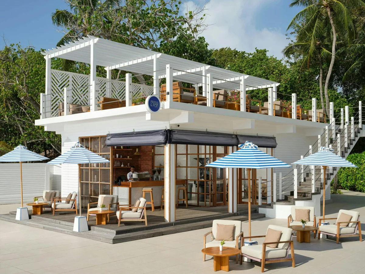 Villa Nautica Paradise Island Resort, dining, North Malé Atoll, Maldives, Press & Pot pavilion: two-story white-clapboard structure with pergola top deck (lounge seating glimpsed above), ground-level outdoor lounge with teak-and-wicker armchairs and blue-and-white striped umbrellas, modern nautical aesthetic in line with the Villa Nautica rebrand.