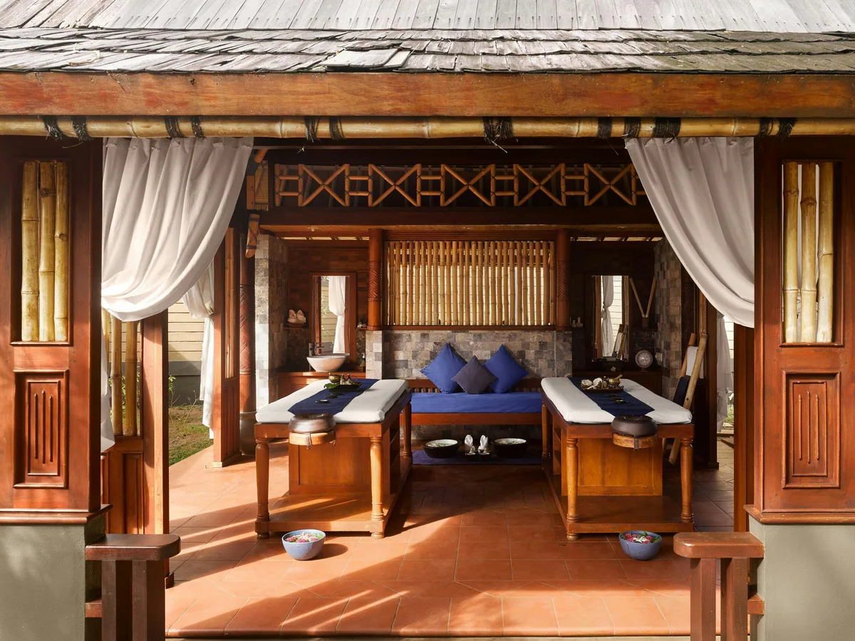 Villa Nautica Paradise Island Resort, spa, North Malé Atoll, Maldives, Araamu Spa treatment pavilion: twin treatment tables under a thatched timber roof with white-curtain side panels drawn back, bamboo screen behind, blue-cushion folded towels on white treatment-table linens, terracotta-tiled floor, carved Maldivian wooden screens at the back wall.