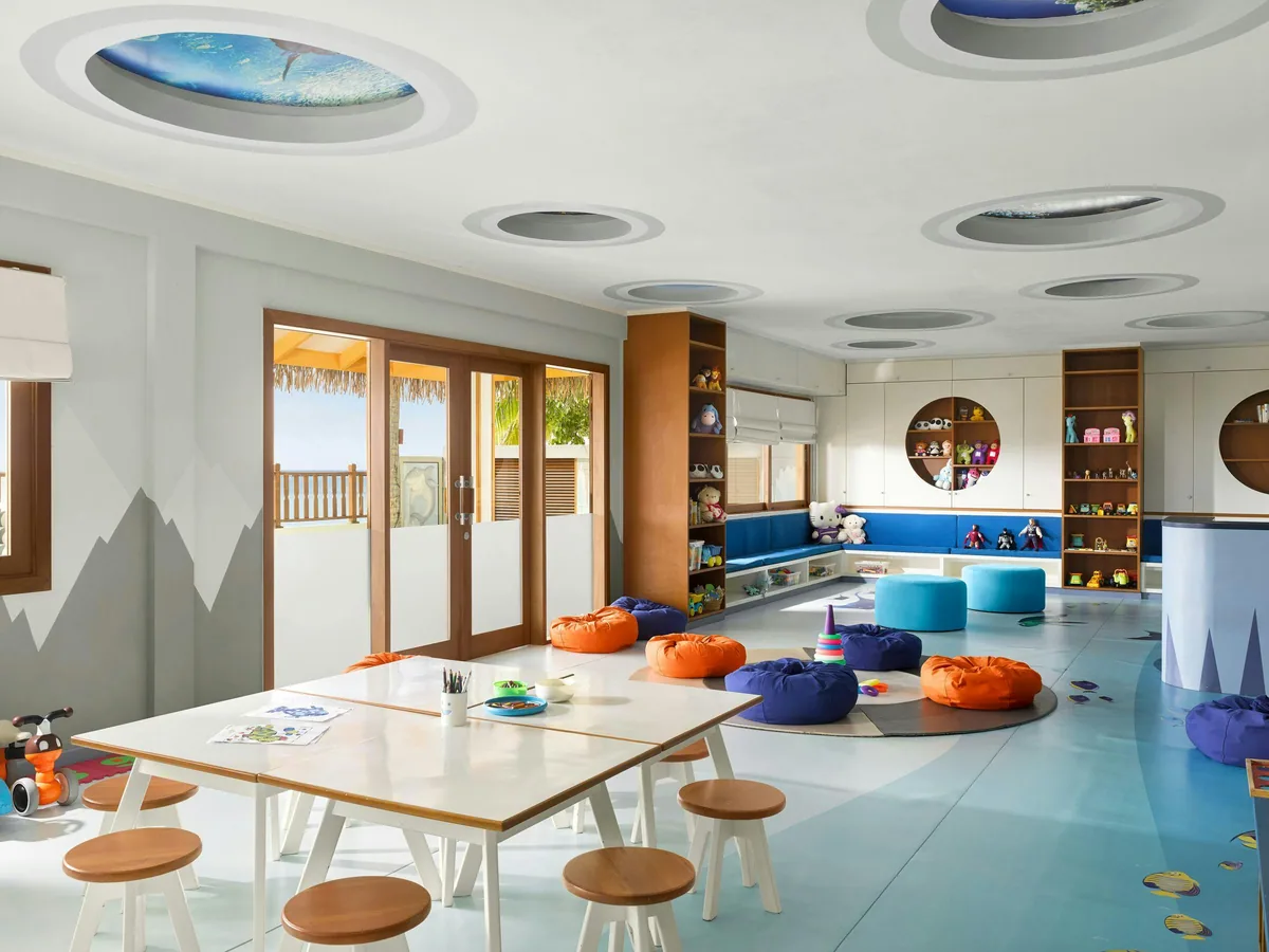 Villa Nautica Paradise Island Resort, activity, North Malé Atoll, Maldives, Wavy Navy Kids Club interior: large play space with porthole-style ceiling cutouts (nautical theme), low children's table with wooden stools, orange and purple beanbag clusters, painted shark fin wall feature, blue floor with painted fish, double doors at the back opening to the beach.
