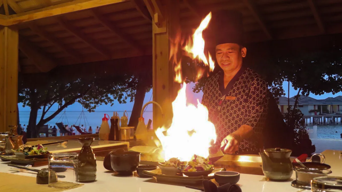Villa Park Sun Island Resort, dining, Alif Dhaal (South Ari) Atoll, Maldives, Sun Teppanyaki chef in branded Japanese-pattern jacket and tall black hat performing flame-grilling at the counter with the dramatic flame plume over the food, sake bottles arrayed, and the twilight overwater villa silhouettes through the open pavilion wall.
