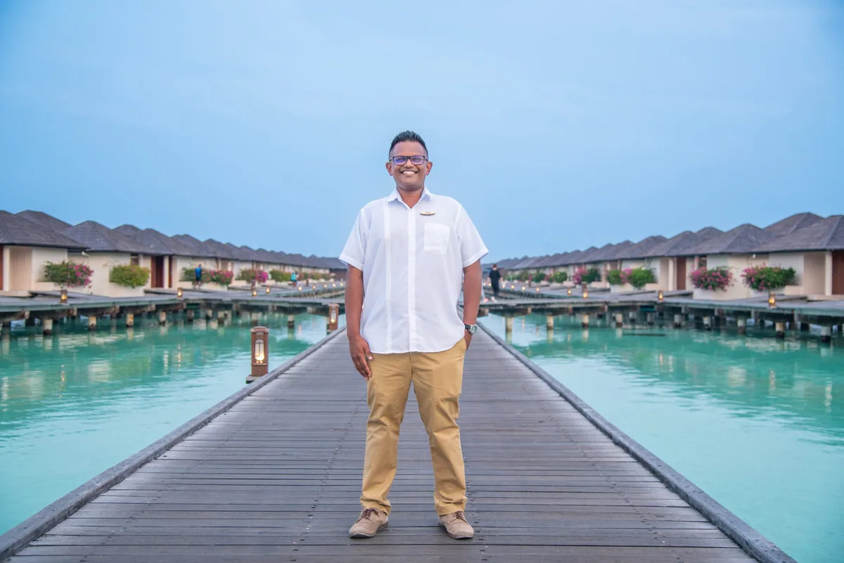 Villa Park Sun Island Resort, setting, Alif Dhaal (South Ari) Atoll, Maldives, Overwater jetty village walk at dusk with the parallel Water Villa rows on both sides, the grey pyramid-tile roofs over white walls, pink bougainvillea boxes between villas, and brass lamps along the timber boardwalk.
