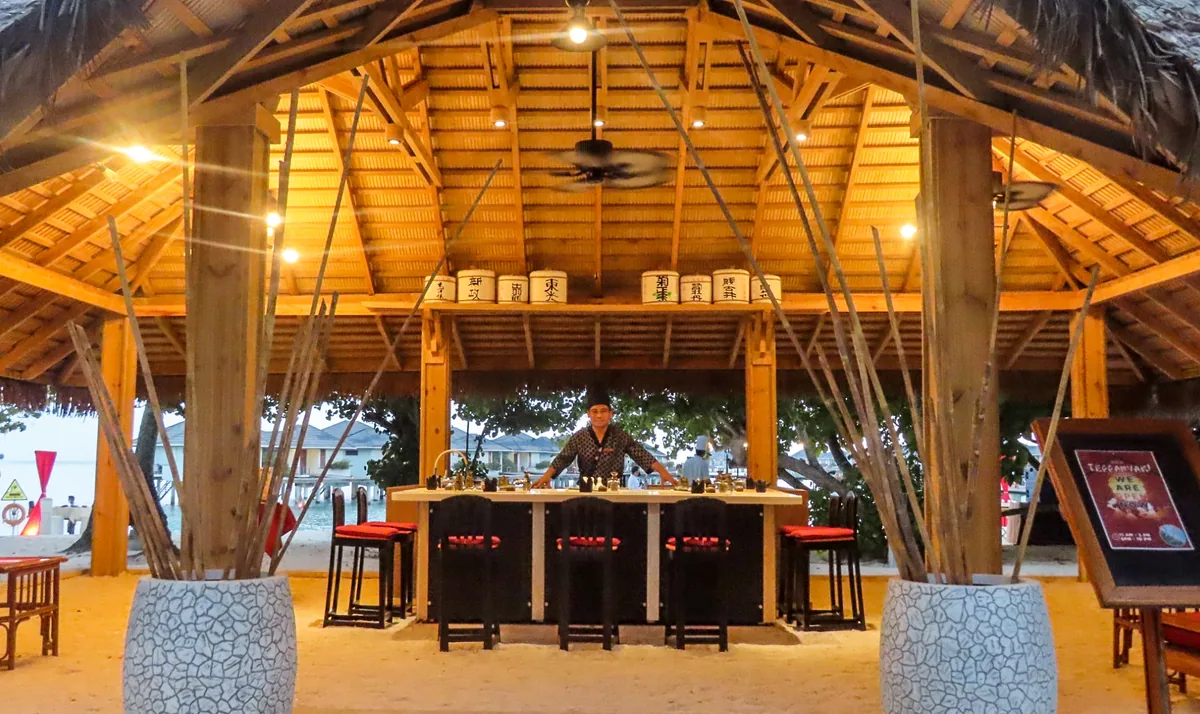 Villa Park Sun Island Resort, dining, Alif Dhaal (South Ari) Atoll, Maldives, Sun Teppanyaki Restaurant pavilion with the bamboo-rib pitched ceiling under thatched gable, the X-frame timber posts, the white sake-barrel cluster suspended at the roof apex, central teppanyaki grill counter with red-cushion stools, and feet-in-sand floor.