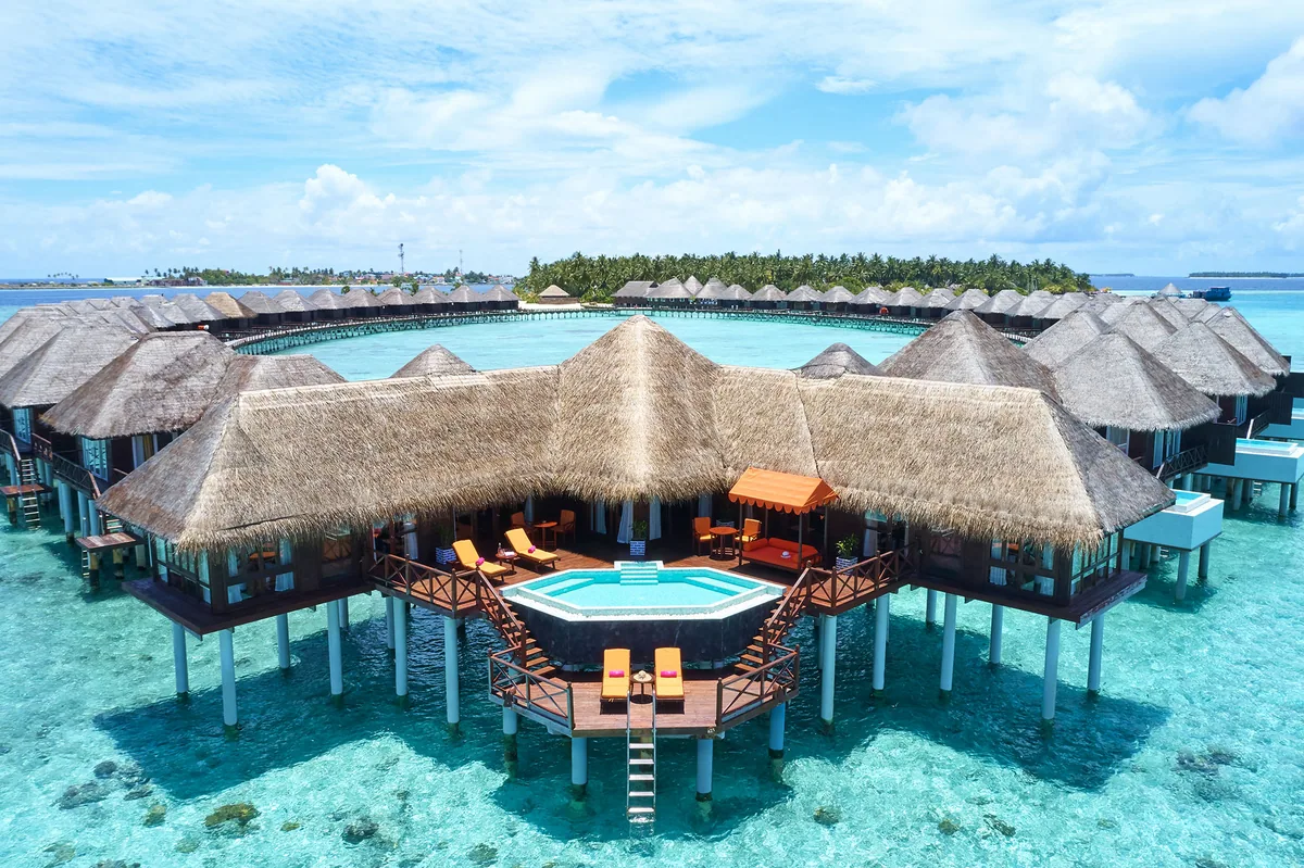 Vilu Reef Beach & Spa Resort, aerial, Dhaalu Atoll, Maldives, Mid-altitude aerial of an over-water villa cluster, central thatched pavilion with circular plunge pool, twin flanking villa wings on stilts, sun loungers and ladder access to the lagoon, palm-canopy main island in the background.