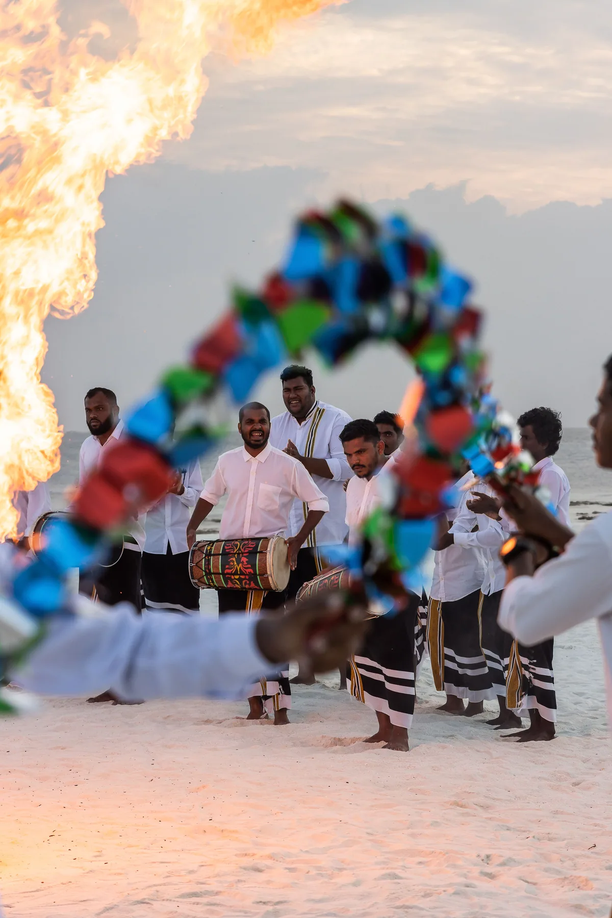 Vilu Reef Beach & Spa Resort, activity, Dhaalu Atoll, Maldives, Traditional Maldivian Bodu Beru drum performance on the beach at sunset, white-shirted drummers in striped sarongs around a barahana drum, a fire performer at left with colorful spinning pom-poms in motion blur in the foreground.