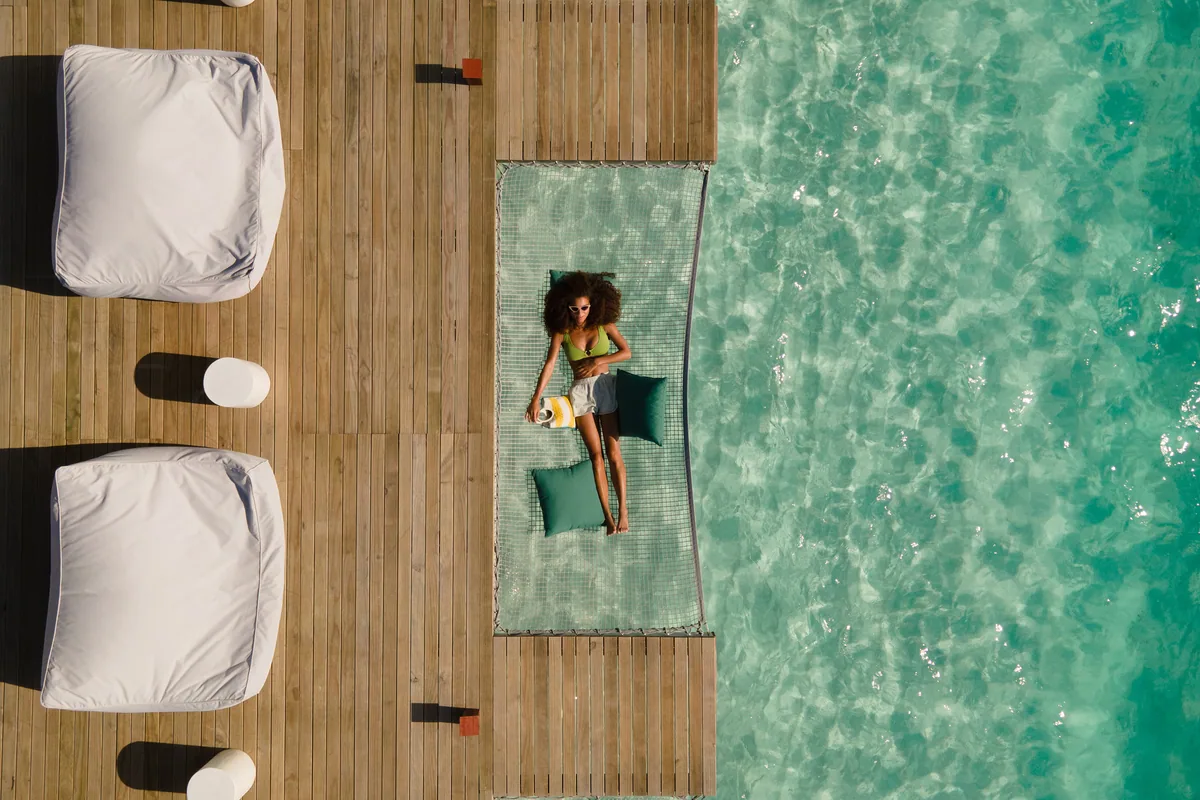 Vilu Reef Beach & Spa Resort, villa overwater, Dhaalu Atoll, Maldives, Top-down view of an over-water villa deck with the property's signature hammock-net inset built into the timber decking, a guest reclining on the net, twin large white bean-bag loungers on the timber boards.