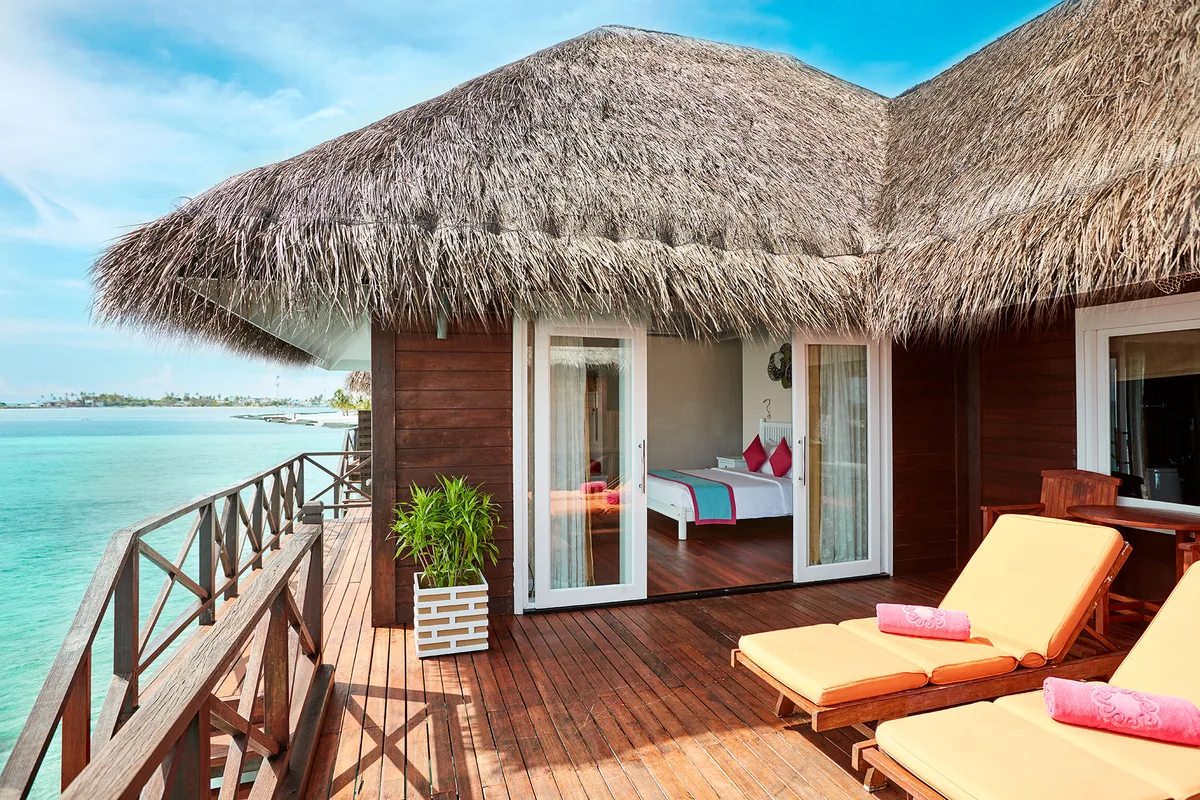 Vilu Reef Beach & Spa Resort, villa overwater, Dhaalu Atoll, Maldives, Reef Villa overwater deck, thatched-roof villa with dark-timber walls and timber-deck balustrade, twin yellow sun loungers with coral-pink towels, glass slider showing villa interior with king bed and pink pop-color pillows, open lagoon and distant reef structures beyond.