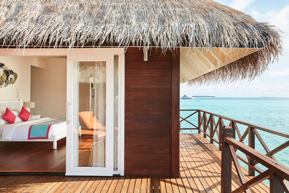 Vilu Reef Beach & Spa Resort, villa overwater, Dhaalu Atoll, Maldives, Sunset Reef Villa entry close-up, the slider opening into a bedroom with white linens and coral-pink and teal pop-color pillows, thatched eave and dark timber wall, balustrade looking out to lagoon with distant island visible at the horizon.