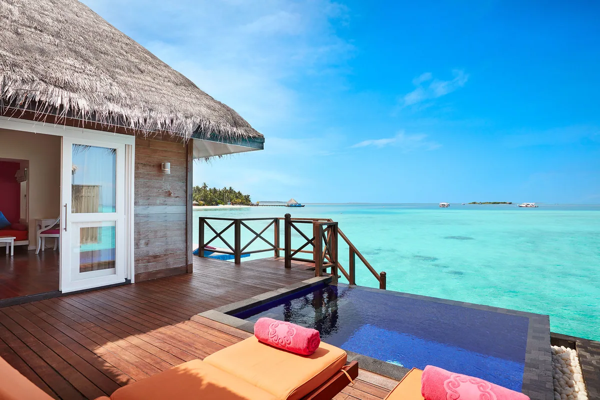 Vilu Reef Beach & Spa Resort, villa overwater, Dhaalu Atoll, Maldives, Aqua Villa with Pool overwater deck, dark-timber walls and thatched roof, a black-tile infinity-edge plunge pool, twin orange sun loungers with coral-pink towel rolls, villa interior visible through glass sliders, open lagoon with distant pavilion and boats on the horizon.