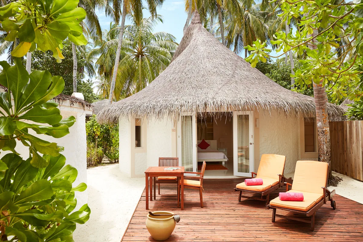 Vilu Reef Beach & Spa Resort, villa beach, Dhaalu Atoll, Maldives, Beach Villa exterior, a round thatched-roof hut with white walls, timber deck with twin yellow sun loungers and coral-pink towels (signature pop-color), a wooden dining set and clay-pot lantern, dense palm canopy framing the entry.