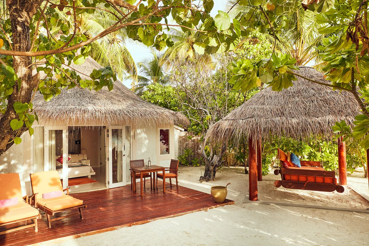 Vilu Reef Beach & Spa Resort, villa beach, Dhaalu Atoll, Maldives, Deluxe Beach Villa with separate daybed pavilion, a round thatched-roof villa at left with sliders into the bedroom and a private deck, a second thatched-roof daybed pavilion at right with rattan swing bench and sun loungers, palm canopy framing both pavilions.