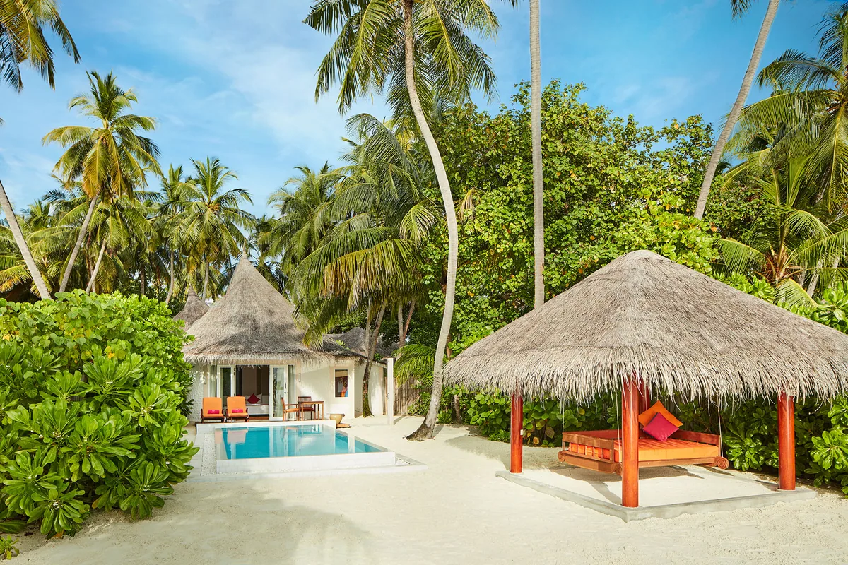 Vilu Reef Beach & Spa Resort, villa beach, Dhaalu Atoll, Maldives, Beach Pool Villa with daybed pavilion, a round thatched-roof villa at left with private rectangular plunge pool, separate thatched-roof daybed pavilion at right with red columns and coral-red mattresses and coral-pink pillows (signature pop-color), palm canopy framing.