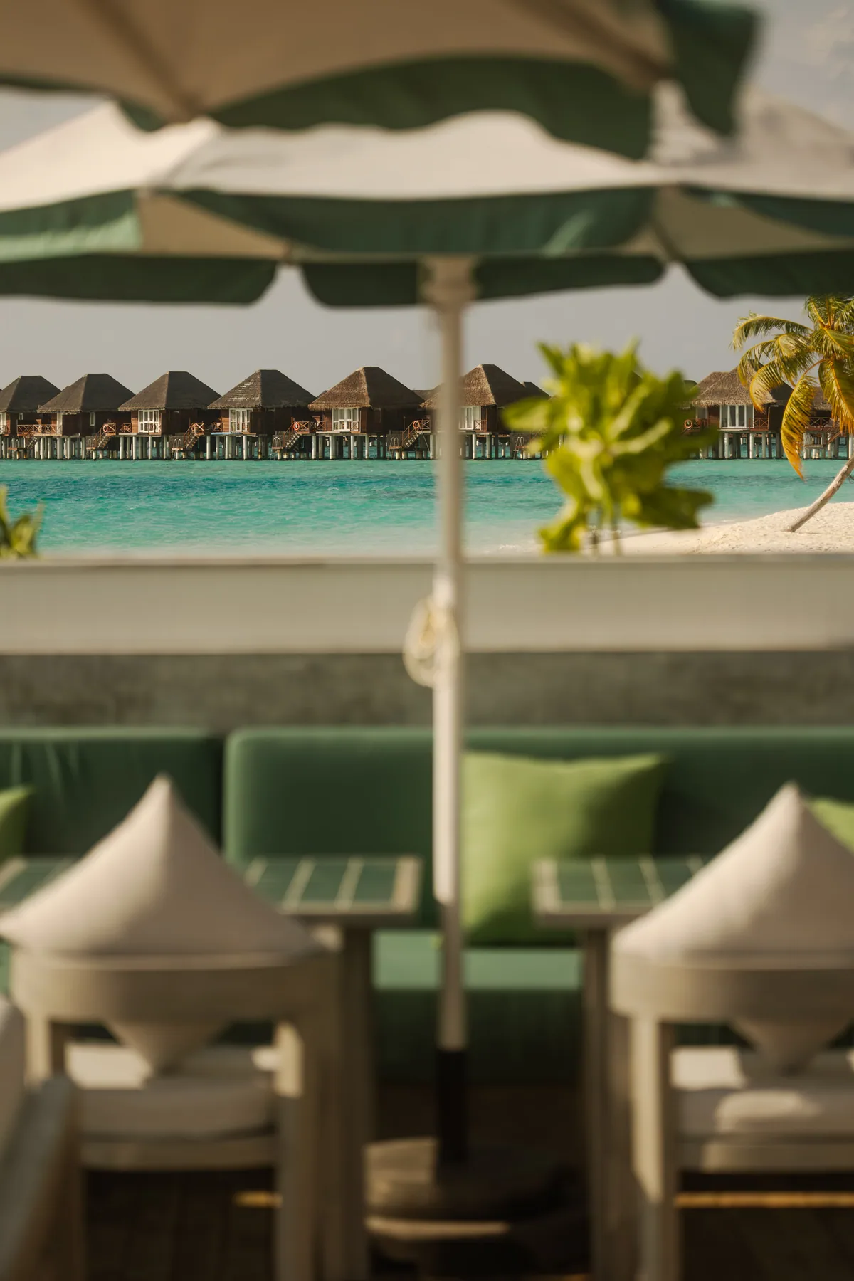 Vilu Reef Beach & Spa Resort, dining, Dhaalu Atoll, Maldives, Beach bar lounge with green-stripe parasols and green-velvet banquette sofas, tepee-style cushion seating in the foreground, the signature over-water villa row visible across the lagoon at the back, palm fronds at right framing.