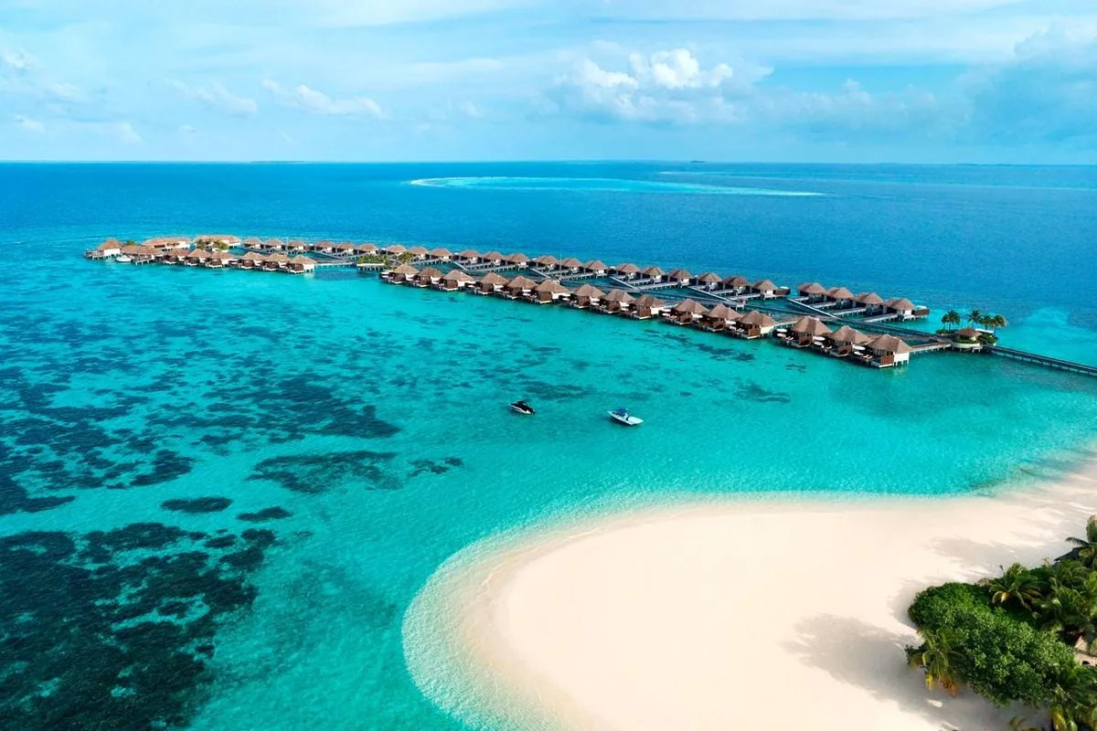 W Maldives, aerial, Alif Dhaal (South Ari) Atoll, Maldives, Fesdu long-axis aerial with the overwater villa row in a straight line atop the deep-blue lagoon shelf, the distinctive curving white-sand spit in foreground, vivid reef pattern under translucent turquoise water, and two excursion boats moored.