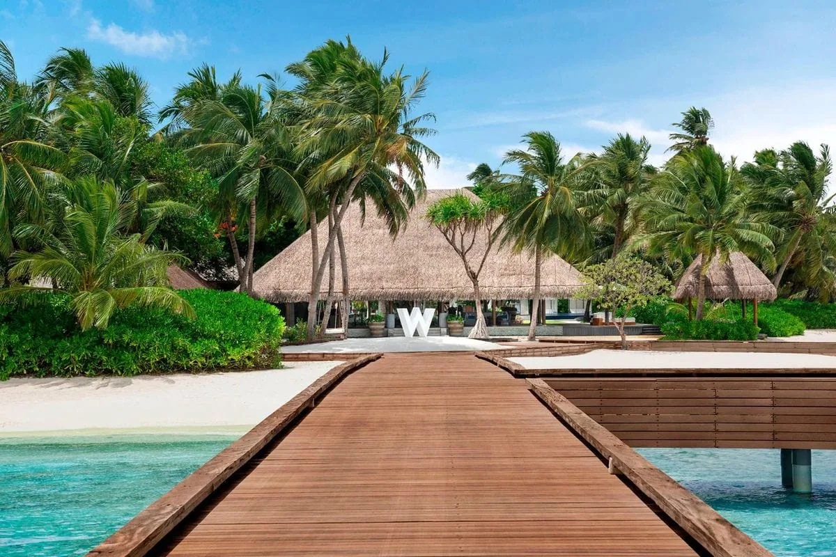 W Maldives, setting, Alif Dhaal (South Ari) Atoll, Maldives, W Maldives arrival with the long timber jetty boardwalk leading to the property entrance, the giant white sculptural W logo at the threshold, thatched-roof entry pavilion behind, and dense palm canopy framing.