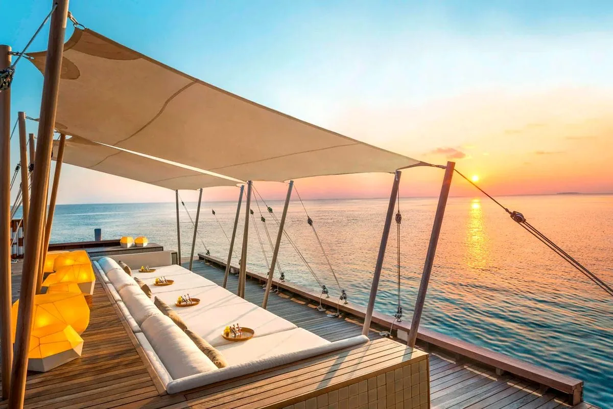 W Maldives, dining, Alif Dhaal (South Ari) Atoll, Maldives, SIP sunset bar with the white-canvas sail-roof shading a triple cushioned daybed run on the timber deck, the glowing yellow modular lantern blocks, the deck balustrade with rigging-rope detail, and the mirror-calm sea at sunset.