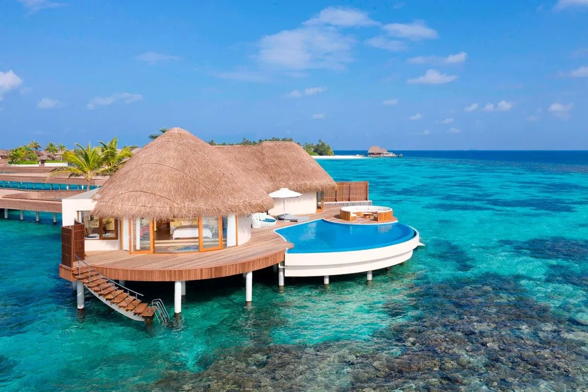 W Maldives, villa overwater, Alif Dhaal (South Ari) Atoll, Maldives, Extreme WOW Ocean Haven, the property's top-tier villa with the thatched-roof curved pavilion on timber stilts, the distinctive wraparound infinity plunge pool with deep-blue tile band, exterior stair into the lagoon, and glowing interior bedroom visible through the retractable glass wall.