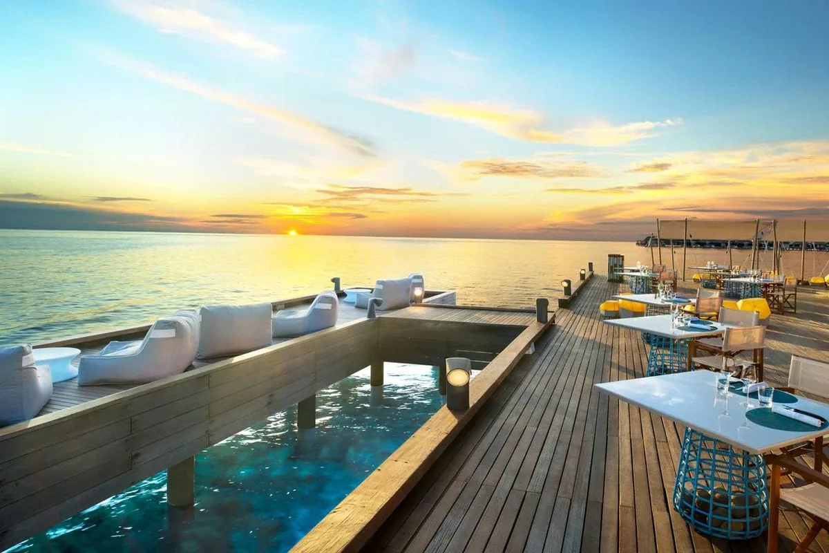W Maldives, dining, Alif Dhaal (South Ari) Atoll, Maldives, SIP sunset deck overview with the sunken sunbed pit ringed with cushion seating around the timber lounge well, the dining-table line with yellow-base wicker chairs, and the deep orange-pink sunset over the open sea.