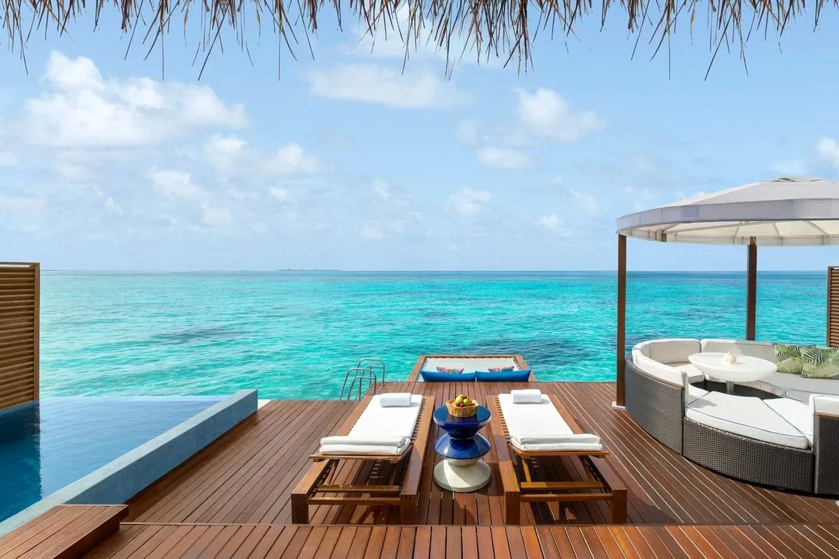 W Maldives, villa overwater, Alif Dhaal (South Ari) Atoll, Maldives, Overwater villa private deck with the wraparound infinity pool, twin wooden loungers with white pads and the cobalt-blue fruit pedestal, the rattan cocoon daybed under the circular white parasol, and the lagoon net hammock at the deck edge.