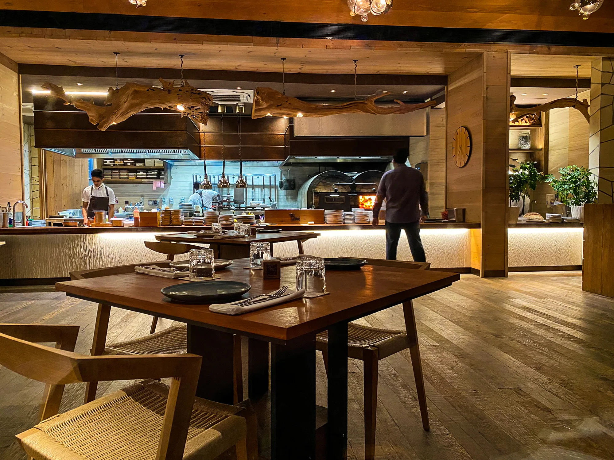 Waldorf Astoria Maldives Ithaafushi, dining, South Malé Atoll, Maldives, The Ledge by Dave Pynt, open kitchen with wood-fired grill in the background, suspended driftwood branch over the counter, hardwood floors and cane chairs around the dining tables.
