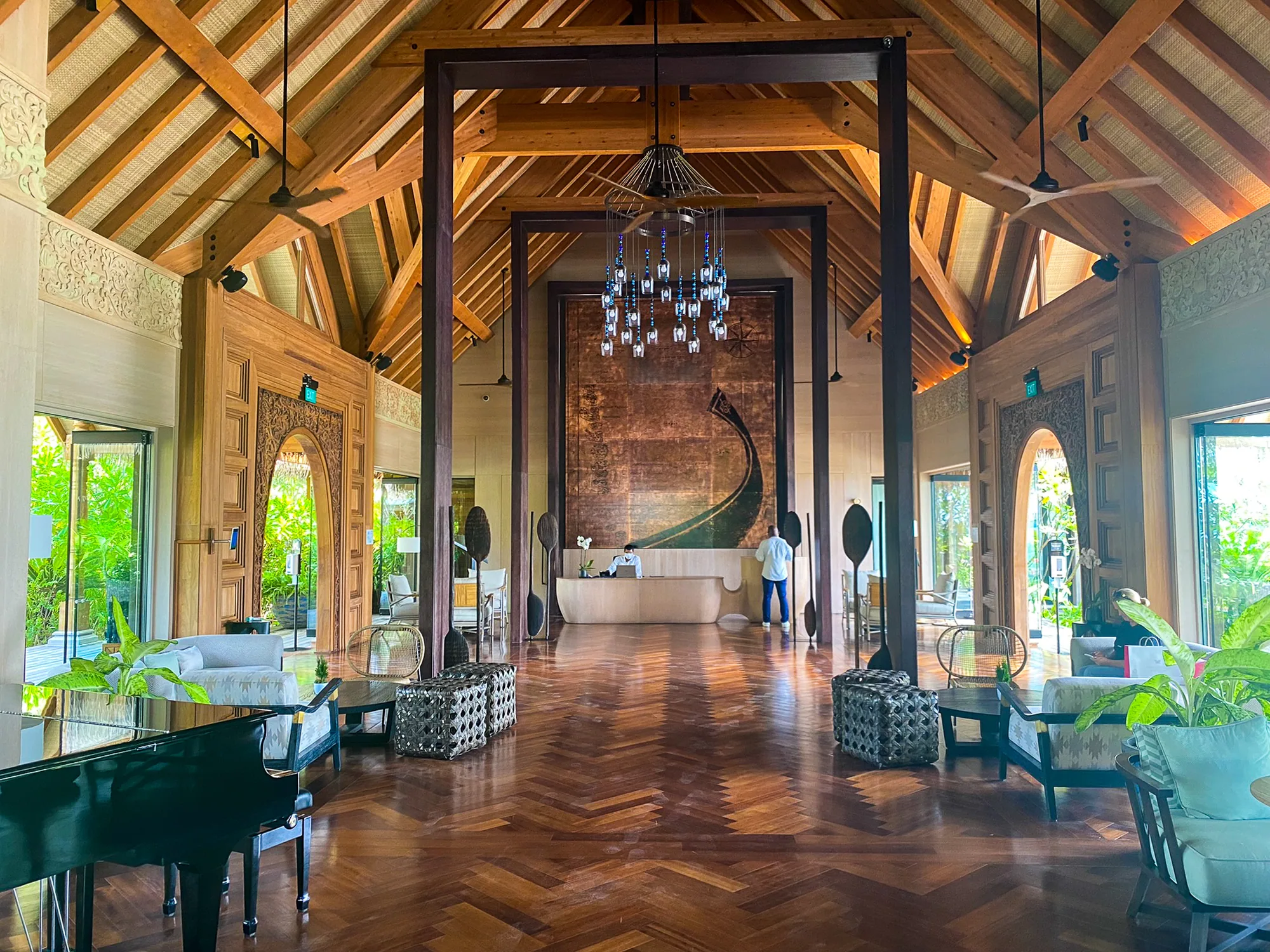 Waldorf Astoria Maldives Ithaafushi, lobby, South Malé Atoll, Maldives, Arrival pavilion interior, exposed-timber pitched roof with feature dhoni-prow wall, beaded chandelier over the reception desk, herringbone parquet floor and a baby grand piano framing the welcome sequence.
