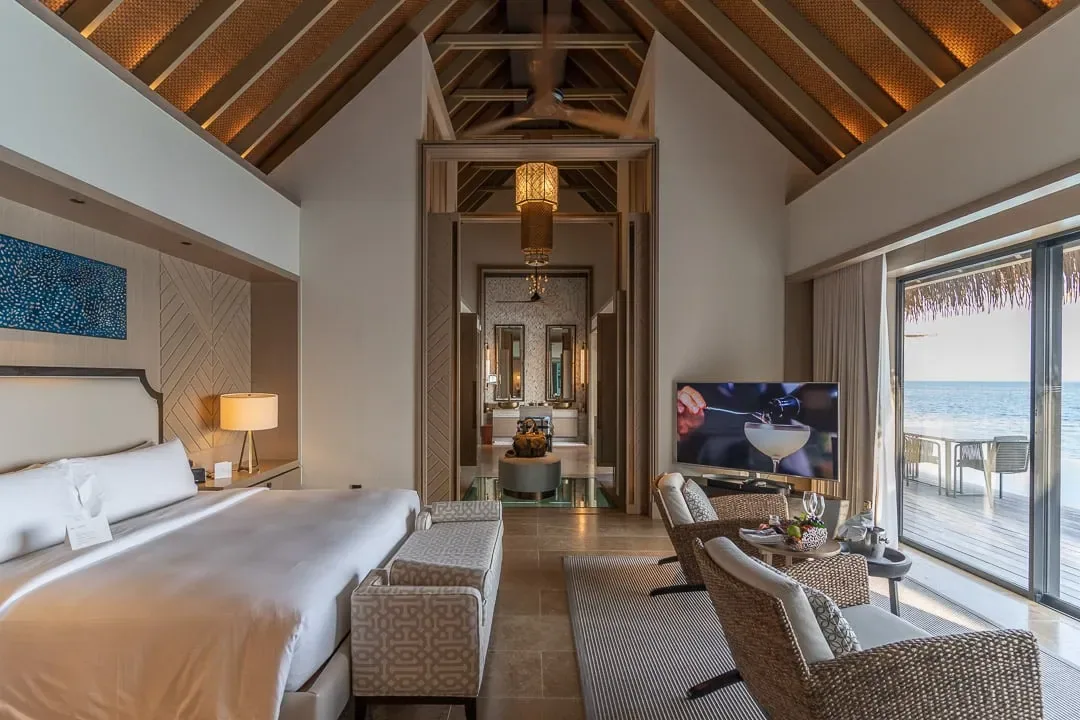 Waldorf Astoria Maldives Ithaafushi, villa overwater, South Malé Atoll, Maldives, Villa interior with cathedral-pitched timber roof, exposed beams under warm uplighting, marine art panel above the king bed, retractable wall opening onto the overwater deck and ocean.