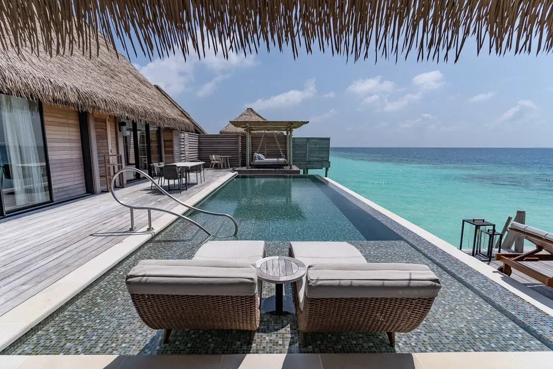 Waldorf Astoria Maldives Ithaafushi, villa overwater, South Malé Atoll, Maldives, Overwater villa exterior, infinity pool extending over the lagoon, rattan loungers on the lower deck, signature thatched roof and dark-wood decking that define the property's overwater inventory.
