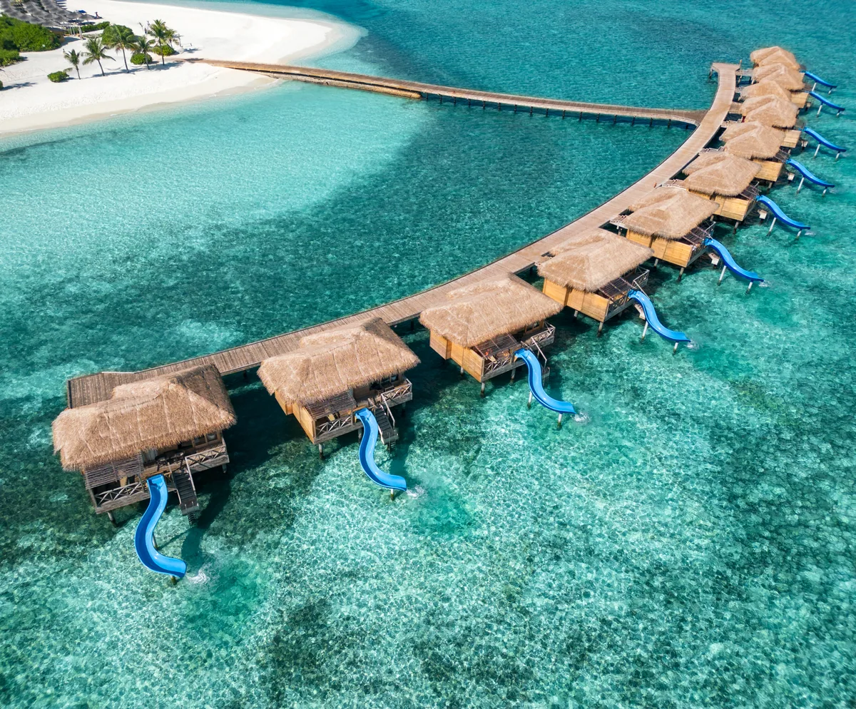 You & Me Maldives by Cocoon Collection, villa overwater, Raa Atoll, Maldives, Aqua Suite with Slide row aerial, curved overwater villa cluster with the iconic bright-blue waterslides projecting from each villa deck down into the turquoise lagoon. The country's only daily-use waterslide-overwater configuration, the property's most-photographed signature feature.